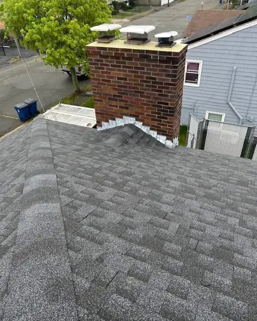 Red brick chimney with three metal flue caps on its crown, rising from a new gray architectural shingle roof with light-colored flashing along the base