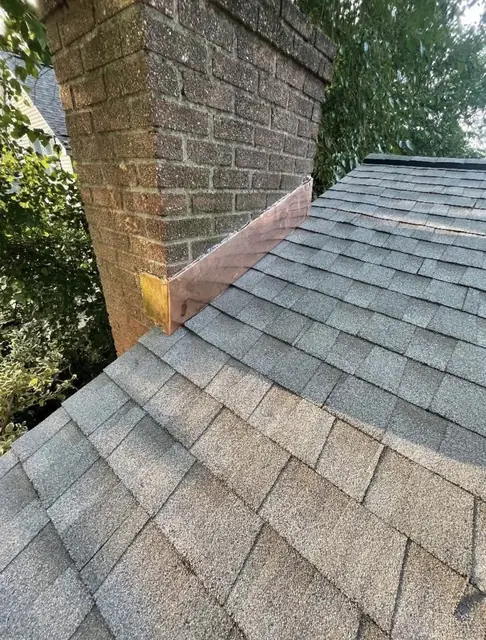 Brick chimney on a gray asphalt shingle roof with new copper flashing running along its side where the brick meets the shingles