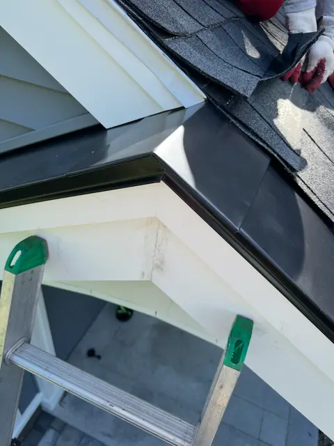 Metal trim and shingle transition at the corner of a porch roof, with a ladder below