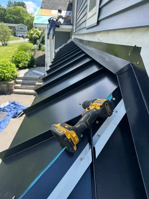 Long run of black standing-seam metal roofing along a house with a drill resting on the seams