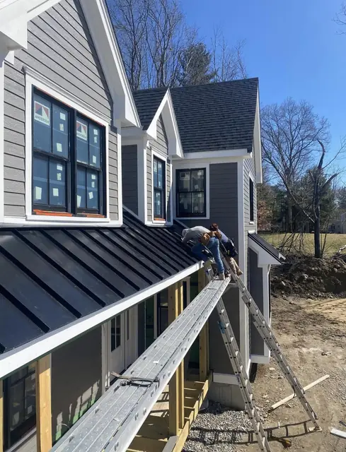Installer on roof staging fastening dark standing-seam panels along the lower edge of a gray house