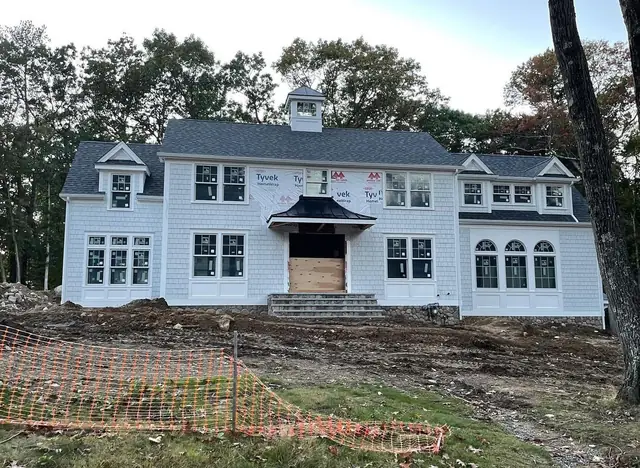 Two-story house with light gray shingle-style siding, white trim and a cupola, with Tyvek housewrap still visible on part of the front wall and a black standing seam metal roof over the front entry