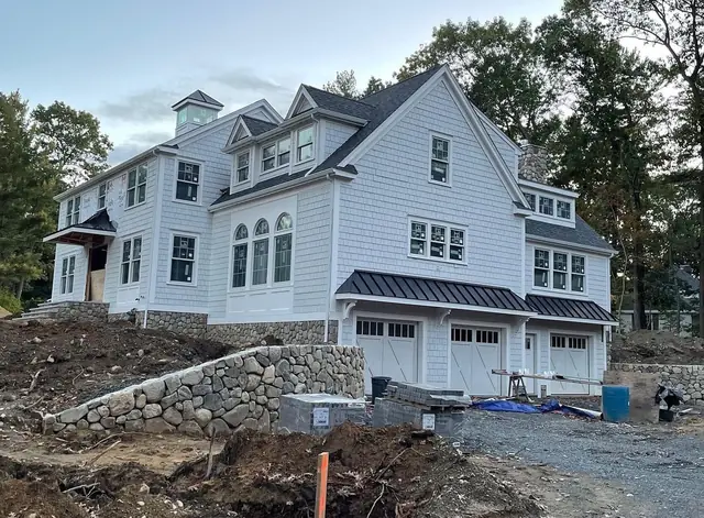 Side view of a gray shingle-style sided home with black standing seam metal roofs over a three-car garage, white carriage-style garage doors and a new fieldstone retaining wall