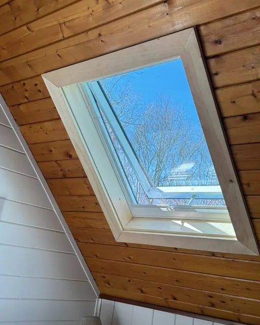 Close-up of a skylight at an angle in a pine plank ceiling with fresh light wood trim, blue sky and tree branches visible through the glass