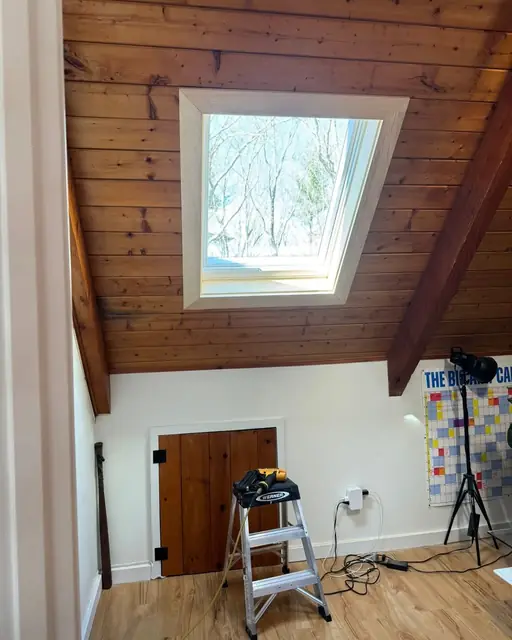 Finished skylight set into a sloped knotty pine plank ceiling with new light wood interior trim, bare winter trees visible through the glass and a stepladder below