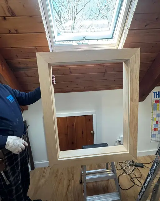 Installer in work gloves holding up a new unfinished wood trim frame beneath a skylight in a pine plank ceiling before fitting it into the opening