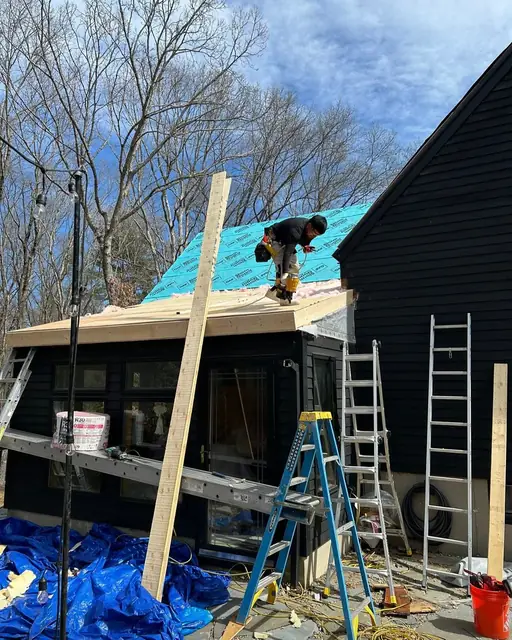 A roofer with a nail gun working at the edge of a low-pitched roof covered in teal synthetic underlayment, with ladders and a scaffold plank set up below