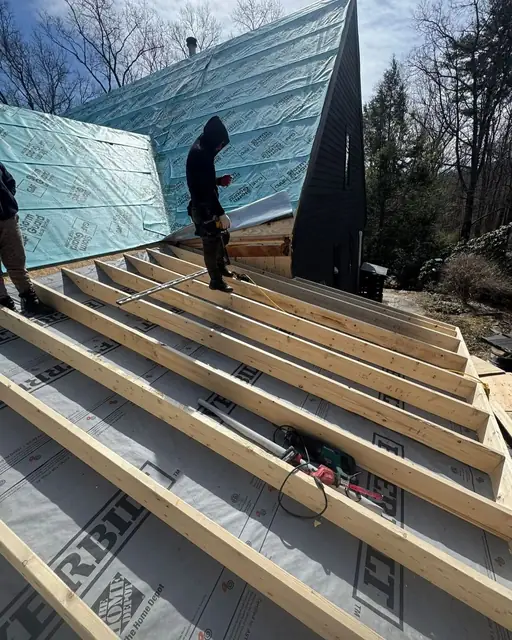 Two workers on a roof deck with new wood strips laid over gray underlayment, next to a steep roof wrapped in teal underlayment