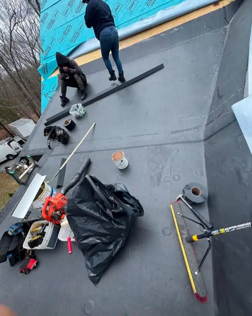 Two roofers working on a low-slope roof covered in a gray membrane, with tools, a leaf blower and a debris bag in the foreground
